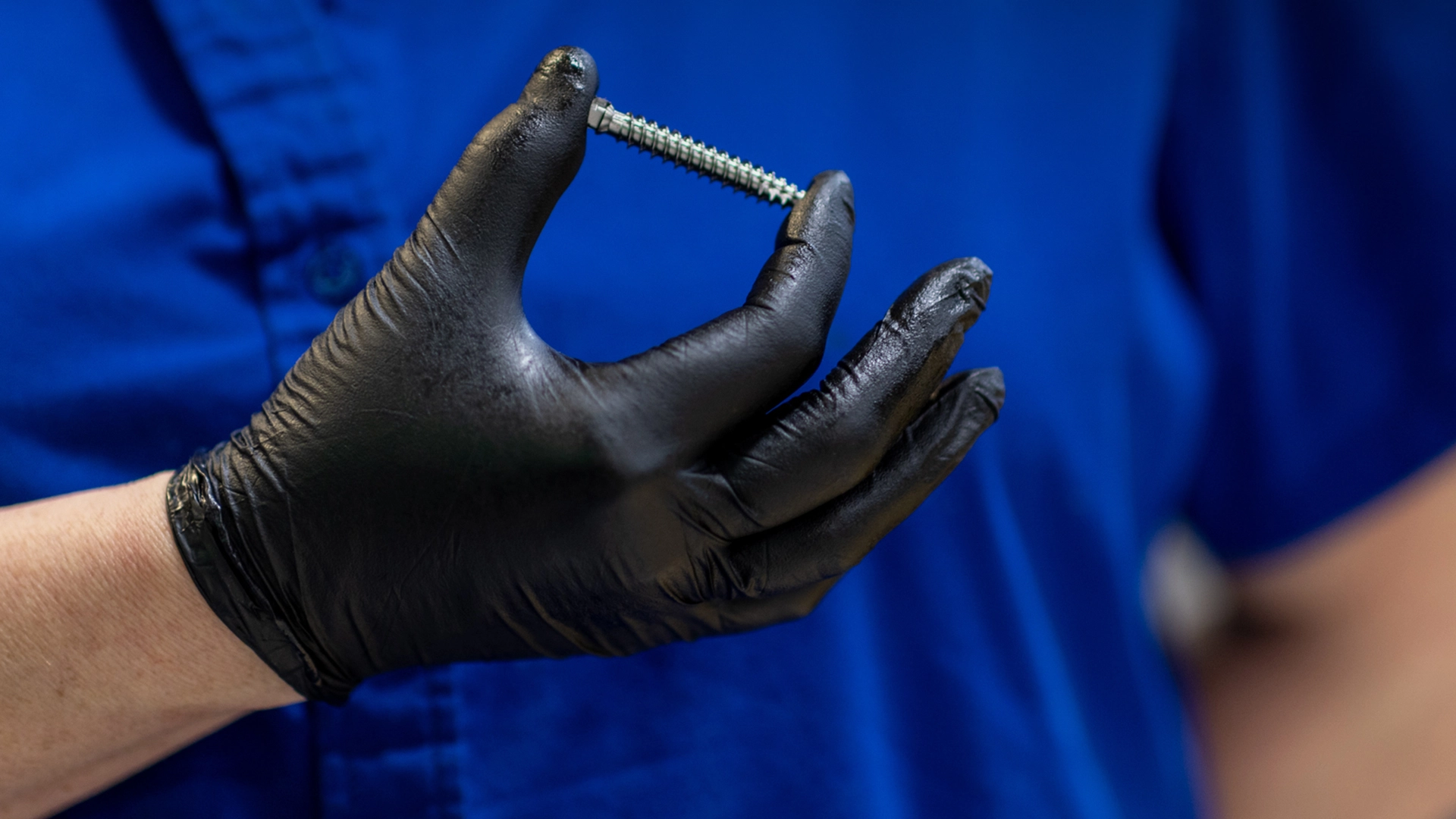 A worker wearing a blue shirt and a black latex glove holds one of the specialty screws that's made at the TOMZ Corporation.